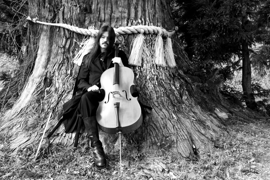 Benjamin Skepper seated with his cello before the sacred cedar of Imaizumi Tenmangu, Rikuzentakata